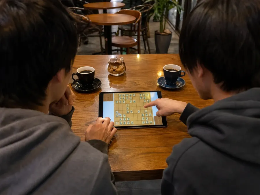 Two people sharing a tablet to play hon-shogi, seen from behind at the shoulders and hands